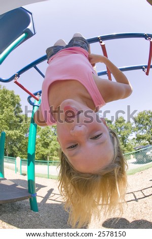 A young blond girl hangs upside down from the monkey bars at a local park.