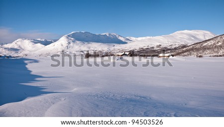 Norwegian winter land with a snow covered lake and a cluster of cabins situated close to the foot of mountains enlightened by the evening sun at easter in the norwegian mountains