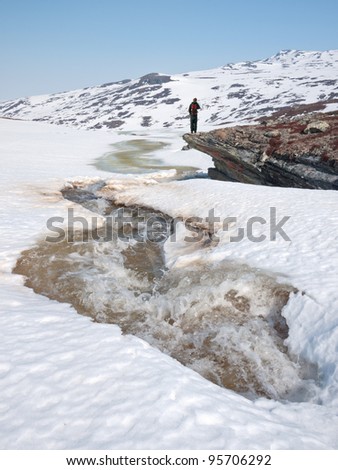 Boy standing on an arrow shaped rock resting during heavy snow melting with ice water floating up and over the snow covered landscape in the norwegian mountains