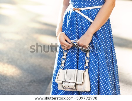 Fashionable woman with white bag and sunglasses in her hands and blue dress