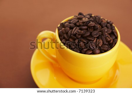 Yellow coffee cup filled with coffee beans over a brown background (shallow dof)