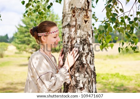 Beautiful young woman in the Russian national dress in a meadow talking to a birch tree.