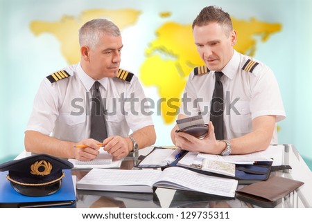 Two airline pilots preparing to flight, checking calculator, papers, flight plan, log book. Pilots are sitting in AIS ARO Air Traffic Services Reporting Office