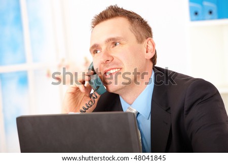 Happy businessman sitting at desk in office, talking on landline phone with laptop and smiling.