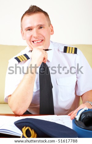 Airline pilot wearing hat, shirt with epaulets and tie filling in and checking papers flight plan, weather forecast. Headset on the table.