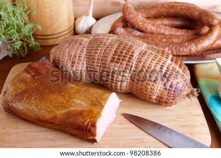 Natural prepared slow food smoked pork sirloin, cured pork shoulder which looks similar to ham and ring-shaped sausage in the background all on the wooden board with herbs