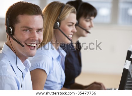 Two female and one male customer service representatives smiling.  They are working on computers and are wearing headsets. The male is looking directly at the camera. Horizontally framed shot.