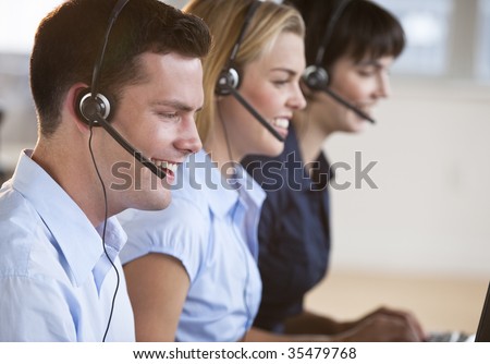 Two female and one male customer service representatives smiling.  They are working on computers and are wearing headsets. Horizontally framed shot.