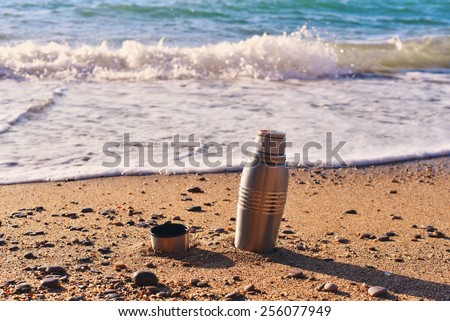 Heat protection-thermos coffee tea cup on the beach, close to the waves, nestled into the center of grainy sand, sandy beach, bank of sea ocean