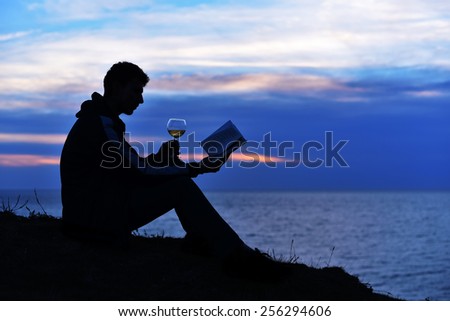 Silhouette guy sitting on breakwater in evening near sea, reads book with glass of wine.\
Silhouette of man reading in the sunset light, sea, ocean, nature, wine