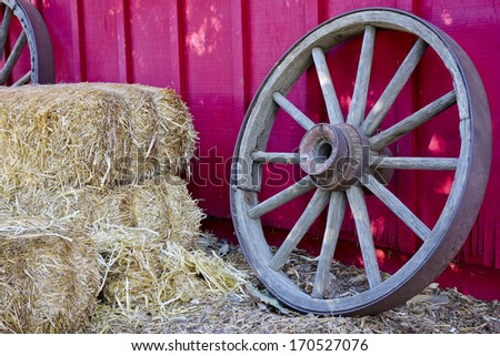 Wooden wheel and stack of hay in next to red ranch house wall