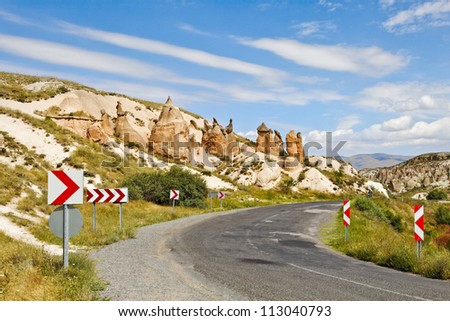 Landscape of Cappadocia rural country lane lined with natural geological volcanic activity, rock formation, limestone, sandstone, chevrons, bend in the road, striking blue sky and clouds.