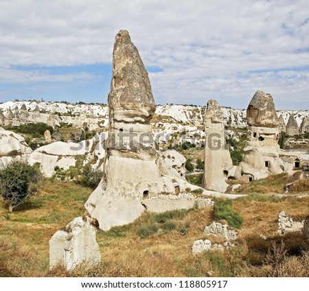 square format group of limestone fairy chimney homes near Goreme Cappadocia Turkey, volcanic lava hills in the background, cloudy blue sky with crop space and copy space