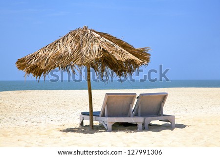 A generic tropical deserted beach with loungers under a coconut leaf parasol. Blue sky, ocean, gentle surf sound and clean sandy beach. Location, Goa India