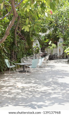 vertical image of shaded patio area with hard standing, tables and wire bucket garden chairs. Mature tree lined hedges and out buildings in distance form a barbecue area