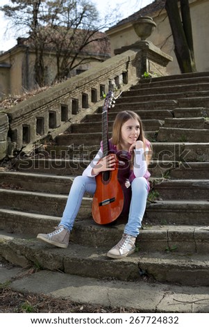 Side View of Beautiful Young Girl With Guitar in Front of Her Sitting on the Foot of the Stairs and Looking Thoughtfully at the Horizon