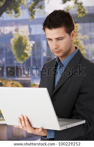 Young businessman standing outside of office with laptop handheld, looking at screen.