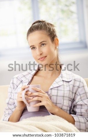 Beautiful woman having morning tea, sitting on sofa with blanket, wearing pyjama, smiling.