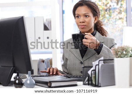 Young afro businesswoman at work in office, drinking tea, using computer.