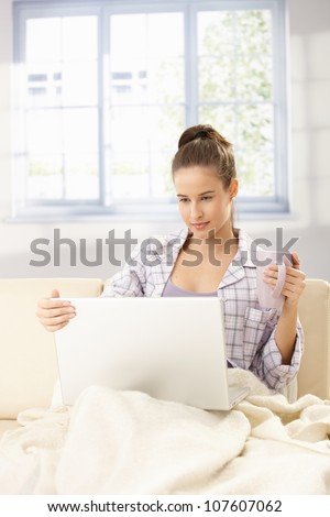 Woman using laptop computer, sitting on sofa in pyjama with blanket, holding coffee mug, smiling in morning light.
