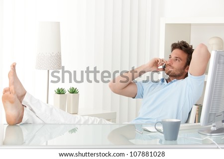 Relaxed man sitting with feet up on desk at home, talking on mobile phone, smiling.