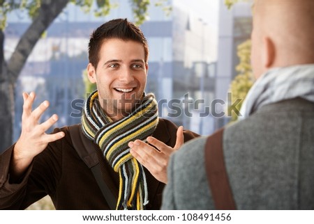 Cheerful guy gesturing to friend in conversation, standing outdoors, wearing scarf, smiling.