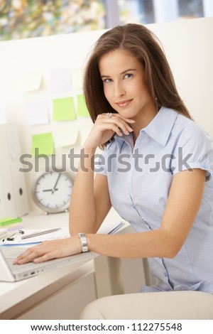 Portrait of female office worker using laptop computer at desk, smiling at camera.