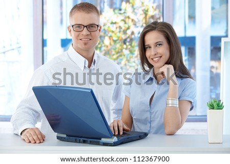 Smiling coworkers standing in office, using laptop computer, looking at camera.