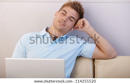 Portrait of boy daydreaming, listening to music with earphones on laptop computer, sitting on sofa with eyes closed.
