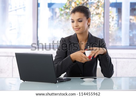 Young woman using laptop in bright office, drinking coffee.