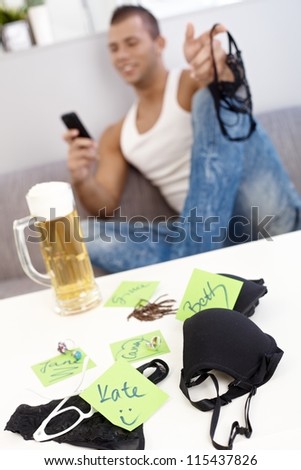 Black female underwear and other accessories matching with girls names, young guy on sofa holding panties and texting, having beer.
