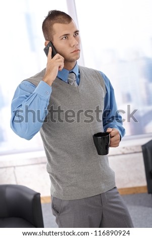 Businessman standing in office with coffee cup handheld, listening to mobile phone conversation, concentrating.