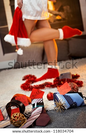 Christmas gifts of tie, belt for men with name tag by pretty girl wearing christmas socks and Santa hat by fireplace.