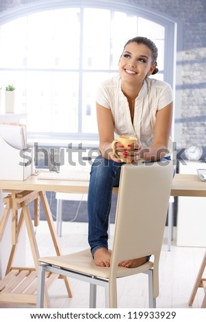 Attractive young girl daydreaming, sitting on top of desk, drinking tea.