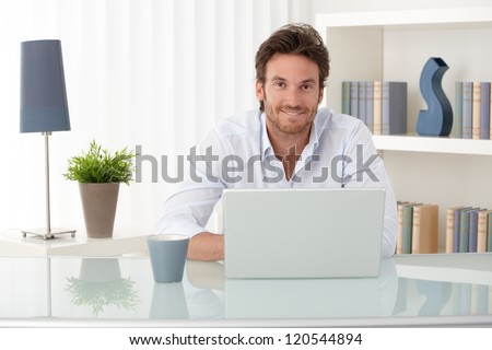 Portrait of goodlooking man sitting at table at home with laptop computer, smiling at camera.