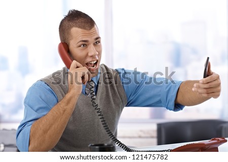 Angry young businessman shouting on landline phone in office, holding mobile phone.
