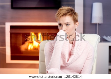 Attractive young woman sitting by fireplace at home in pink pullover, thinking.