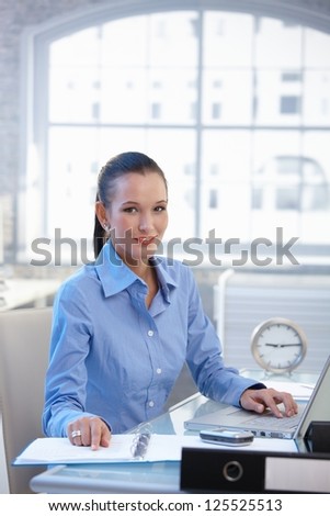 Smiling office worker girl busy at desk with laptop computer and documents.