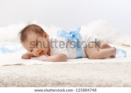 Newborn baby lying on front in white bodysuit and blue ribbon around waist, lifting head.