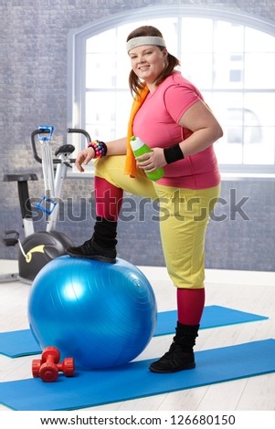 Young fat woman taking a break at the gym, resting leg on fit ball, drinking water.