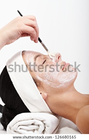 Young woman getting beauty treatment, beautician applying cream on her face, side view.