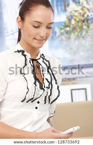 Young smiling businesswoman in fancy shirt texting on mobile phone in office, looking down.