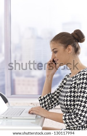 Side view of attractive young businesswoman sitting at table, using laptop computer, talking on mobile phone.