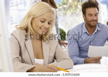 Attractive blonde businesswoman sitting at desk in office, writing notes, smiling.
