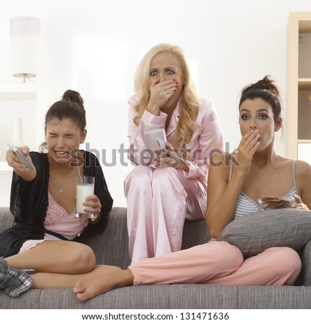 Girls watching thriller on tv at home in pyjamas, looking with horror on face.