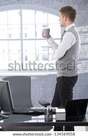 Businessman standing in bright office, looking out of window, drinking tea, thinking.