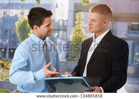 Businessman explaining colleague computer work on laptop, standing outside of office building.