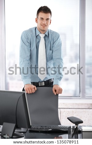 Young manager standing in office behind chair, smiling at camera.