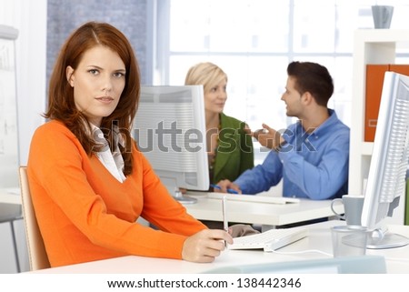 Portrait of office worker woman sitting at desk, looking at camera, colleagues at background.