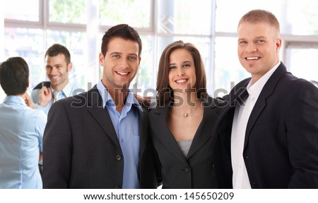 Portrait of successful young business team standing at bright office, smiling happy, looking at camera.